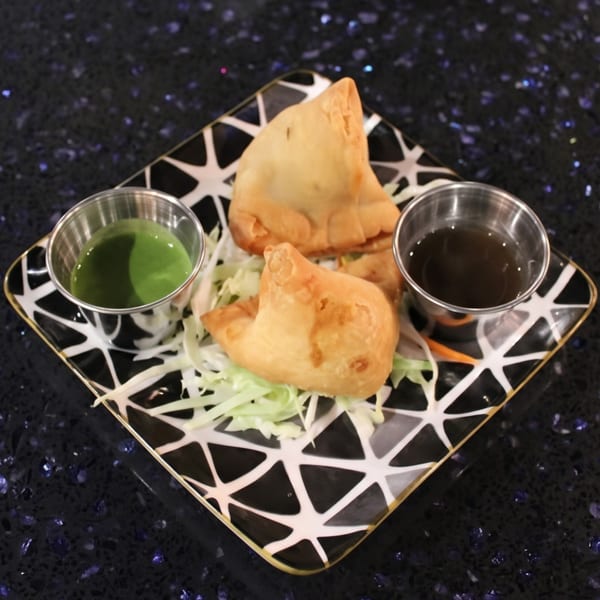 Three golden-brown samosas on a bed of shredded cabbage, with green and brown dipping sauces.
